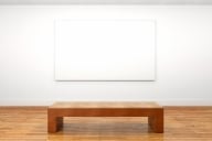 An empty wooden museum bench sits in front of a blank white canvas on a museum wall.