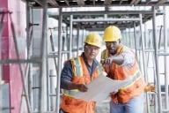 Two men in hard hats and high-visibility vests lean over a blueprint, one pointing. 