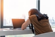 A young woman sits at her desk with her head in her hands facing a dark laptop screen