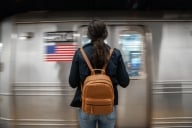 Individual waits on the platform, with a backpack style pocketbook, waiting for the train to stop. 