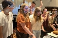 A group of students hold and play musical instruments in a classroom, smiling. 