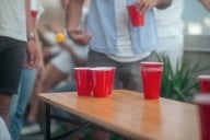 Young people bounce orange ball over red cups in a game of beer pong.