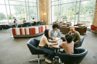 Four college students work at a round table with three other students in the background at the library.