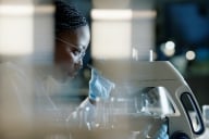 A scientist wearing protective eyewear and gloves looks into a microscope in a lab.