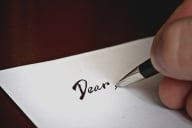 A close-up of a hand holding a pen to paper, having written the word "Dear," as if to open a letter.