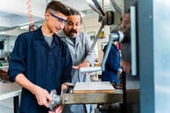 A young man operates a machine wearing safety goggles with an instructor behind him. 