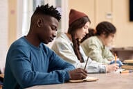 Side view portrait of young African American man writing in notebook sitting at table with group of students in row during college class copy space