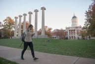 A student walks down a path on a college campus in front of a row of columns