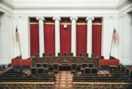 A photo of the interior of the U.S. Supreme Court chambers, showing a curved table with nine chairs in front of a set of five long red curtains hanging between four white columns.