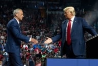 A photo of Robert F. Kennedy Jr. walking toward Donald Trump on stage at a rally. Both men have hands outstretched for a handshake.