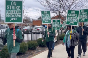 Demonstrators walk on a sidewalk next to a parking lot holding green signs that say "On Strike For a Fair Contract"