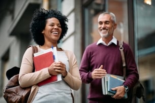 A woman and man, older than traditional college students, laugh while carrying books and wearing backpacks. 