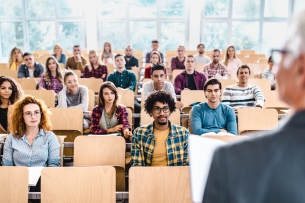 A professor stands in front of a classroom of students with some of the chairs unfilled.  