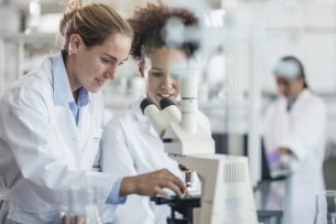 Female scientists in white coats with their hair pulled back stand beside a microscope.