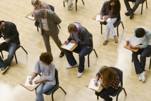 Students sit in a classroom taking a test as a teacher walks by.