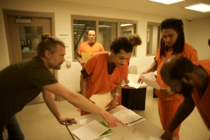 Nate Johnson, founder of FreeWriters, and three incarcerated students in orange uniforms stand around a table covered in papers.