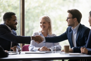 Four diverse people sit around a table, while one man stretches out a hand to welcome another