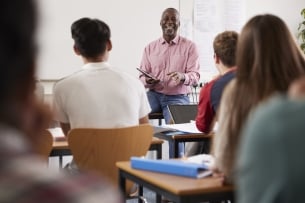 A male teacher sits on a desk as he instructs class.