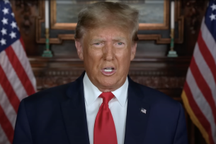 Donald Trump, wearing a blue suit and red tie, speaks in front of two American flags.