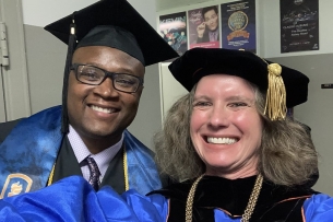 A Black male student in a graduation gown and mortarboard stands next to a light-skinned woman with gray hair who is also wearing academic regalia.