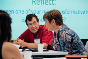 Attendees of a conference, two middle-aged light-skinned people, talk at a table. 