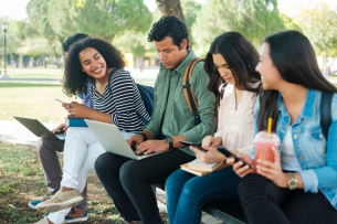 Five students sit outside on a college campus on their laptops and phones.