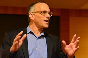 A man in a blazer and glasses talks using his hands.