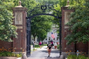 A student with a backpack walks under the entryway to a tree-lined college campus.