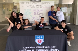 Several students and several more library staff members sitting at a table outside the library at Loyola Marymount University in California to welcome students to the Long Night Against Procrastination event. A whiteboard behind them details the event. Most people in the photo are in humorous poses or making funny faces.