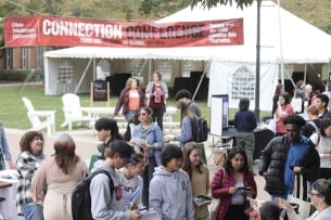 Students gather in small groups at at tables on the grounds of Ohio Wesleyan University.  A banner saying "Connection Conference" hangs in the background.  