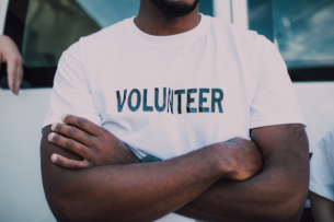 A man stands with his arms crossed wearing a white T-shirt that says "VOLUNTEER." Only the man's chest and arms are visible in the picture.