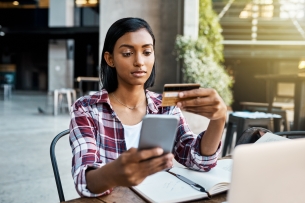 A young woman in a red flannel holds up her credit card as she makes a purchase on her cellphone.