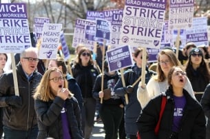 A photograph of striking University of Michigan graduate student workers, holding purple and white strike signs.