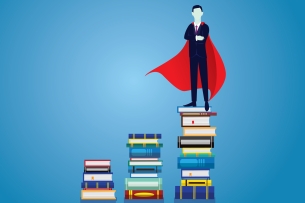 Man in a suit and a superman cape standing on a stack of books with two other stacks to the side of him