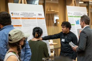 Two students in a large room share their science research poster with a professor as others walk by.