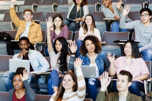 A group of students in a lecture hall raises their hands.
