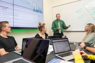 Three students sit at a desk with open laptops while the instructor stands at the whiteboard, talking.