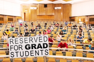 A lecture hall of students with two empty rows of chairs at the bottom and a sign that says "reserved for grad students."