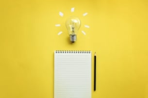 Pad of paper and pencil on a yellow background with a bright light bulb above them