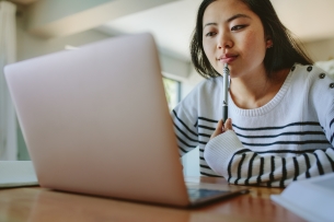 Asian woman studying at home using laptop on the table, holding a pen in hand.