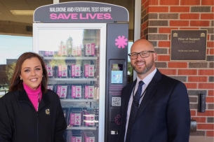 Two individuals stand next to a pink-and-black vending machine containing Narcan.