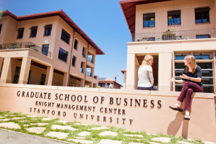 Two students sit on a Stanford Graduate School of Business sign on the Palo Alto campus.