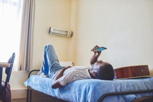 Tired young man lies on his bed while texting on his cellphone inside of his apartment during the day