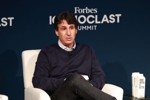 Marc Rowan, a white man with dark hair, seated, in front of a blue wall that bears the name of the event, the Forbes Iconoclast Summit.