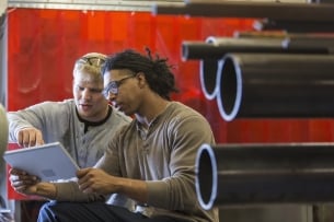 One man shows another man a digital tablet. There are pipes in the background.