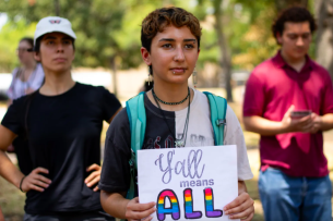 A student holds a sign reading "y'all means all," with "all" in rainbow colors.