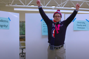 A man stands in front of whiteboards with signs taped to them reading "academic integrity" and "diversity, equity and inclusion" with both hands raised excitedly.