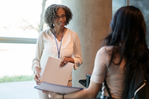 A female professor, standing and facing the camera, listens to a female undergraduate student talking to her.