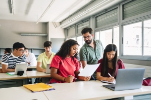 A classroom full of students sit in rows. One student shows a paper to a professor. 