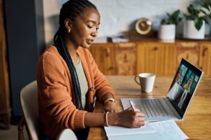 Woman sits in front of laptop and Zoom conference in home office, writing notes and listening to colleagues during online meeting.